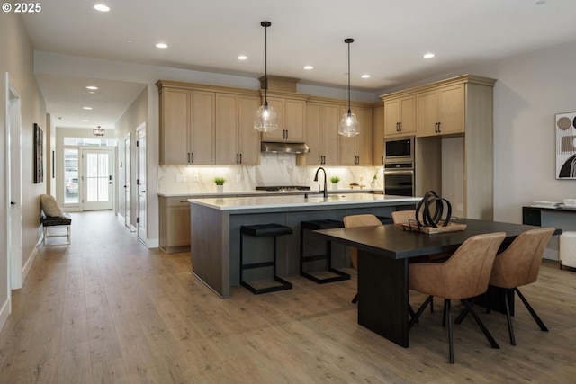 kitchen with light brown cabinets, light wood finished floors, decorative backsplash, under cabinet range hood, and appliances with stainless steel finishes