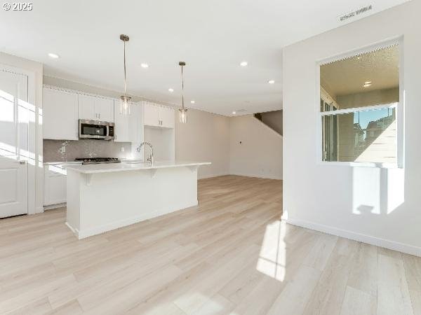 kitchen with white cabinets, light countertops, stainless steel microwave, a center island with sink, and pendant lighting