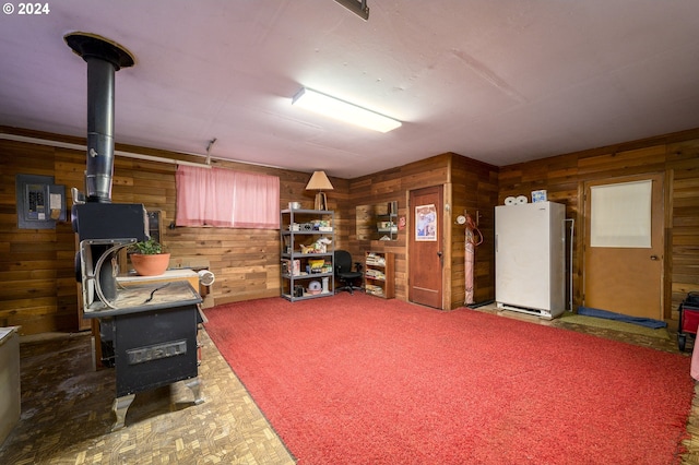 basement featuring a wood stove, wooden walls, electric panel, and white refrigerator