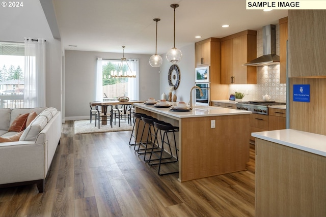 kitchen with backsplash, an island with sink, a chandelier, wall chimney exhaust hood, and hardwood / wood-style flooring