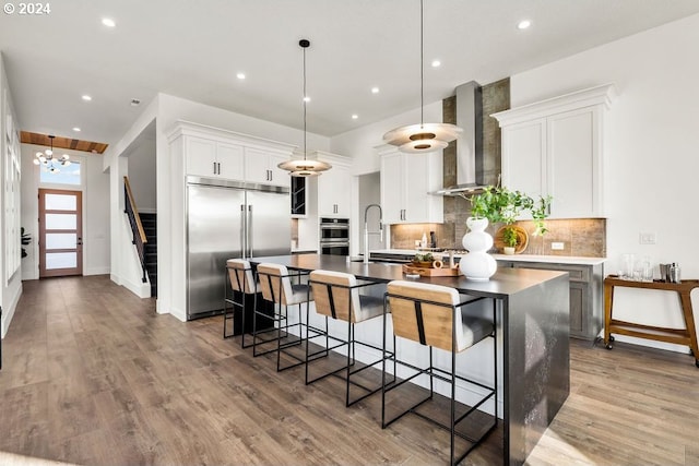 kitchen with a kitchen island with sink, white cabinets, wall chimney exhaust hood, decorative light fixtures, and stainless steel appliances