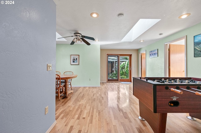 recreation room with a skylight, ceiling fan, a textured wall, baseboards, and light wood-type flooring