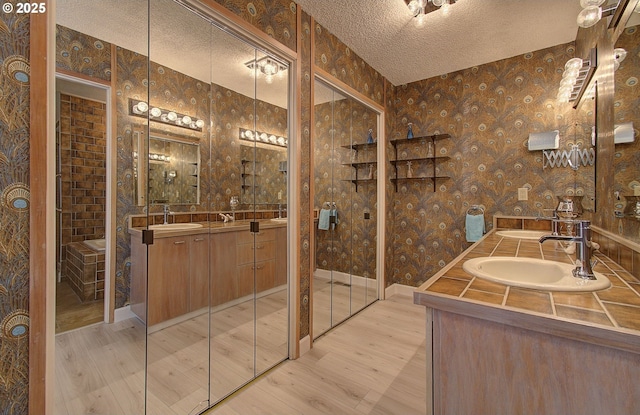 bathroom featuring wood finished floors, a textured ceiling, wallpapered walls, and double vanity