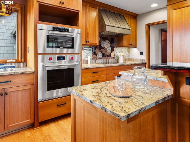 kitchen with stainless steel double oven, wall chimney range hood, light stone counters, decorative backsplash, and black electric stovetop