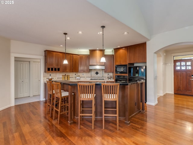 kitchen featuring arched walkways, tasteful backsplash, under cabinet range hood, an island with sink, and black appliances