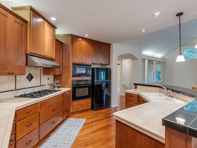 kitchen featuring brown cabinets, black appliances, under cabinet range hood, a sink, and arched walkways