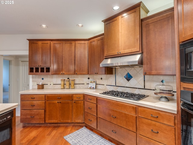 kitchen with light wood-type flooring, black appliances, under cabinet range hood, backsplash, and brown cabinetry