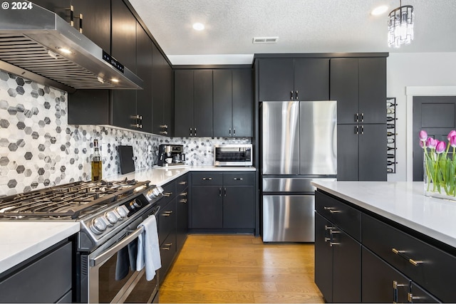 kitchen with backsplash, exhaust hood, light hardwood / wood-style flooring, appliances with stainless steel finishes, and decorative light fixtures