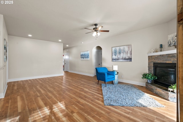 unfurnished room with arched walkways, a stone fireplace, a ceiling fan, and wood finished floors