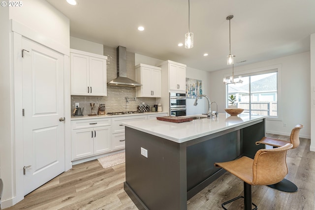 kitchen with an island with sink, pendant lighting, white cabinets, and wall chimney exhaust hood