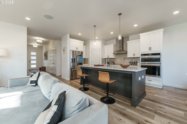 kitchen with hanging light fixtures, stainless steel appliances, an island with sink, white cabinets, and wall chimney exhaust hood