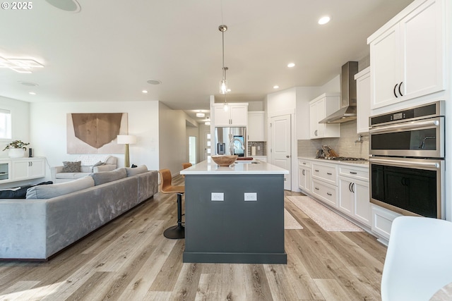 kitchen featuring wall chimney range hood, an island with sink, pendant lighting, stainless steel appliances, and white cabinets