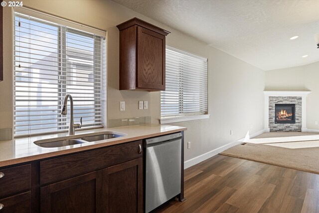 kitchen with dark brown cabinetry, sink, stainless steel dishwasher, a stone fireplace, and dark hardwood / wood-style flooring