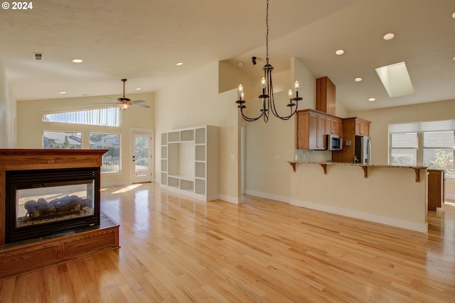 living room with a skylight, high vaulted ceiling, light hardwood / wood-style floors, and ceiling fan with notable chandelier