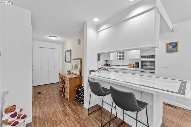 kitchen with stainless steel oven, a breakfast bar, tile countertops, wood finished floors, and white cabinetry