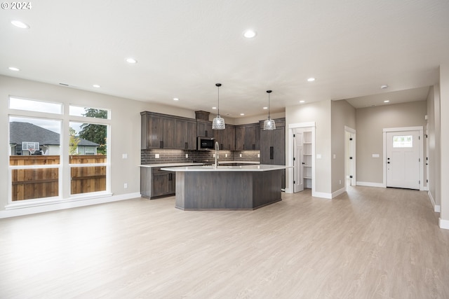 kitchen with light wood-type flooring, backsplash, dark brown cabinets, a kitchen island with sink, and hanging light fixtures