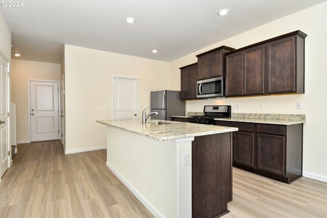 kitchen with light wood finished floors, a kitchen island with sink, dark brown cabinets, stainless steel appliances, and a sink