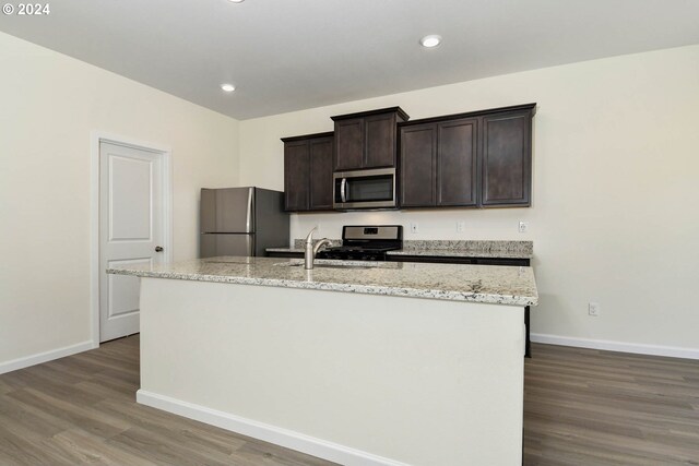 kitchen with a center island with sink, wood finished floors, stainless steel appliances, baseboards, and dark brown cabinets