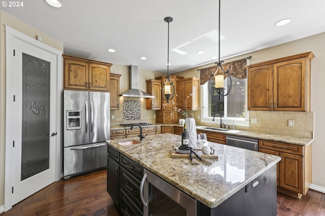 kitchen with stainless steel appliances, sink, wall chimney range hood, decorative light fixtures, and a center island with sink