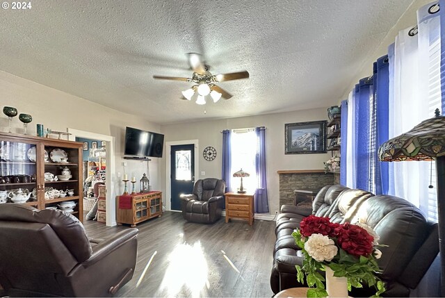 living room with a textured ceiling, dark hardwood / wood-style floors, a stone fireplace, and ceiling fan