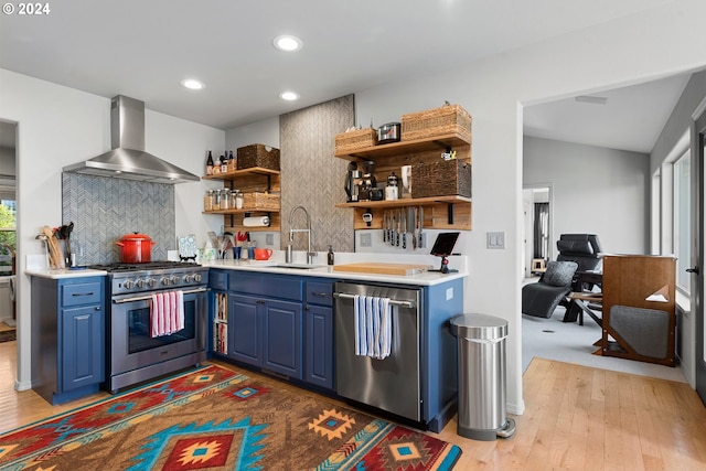 kitchen with stainless steel appliances, a sink, wall chimney range hood, and blue cabinetry