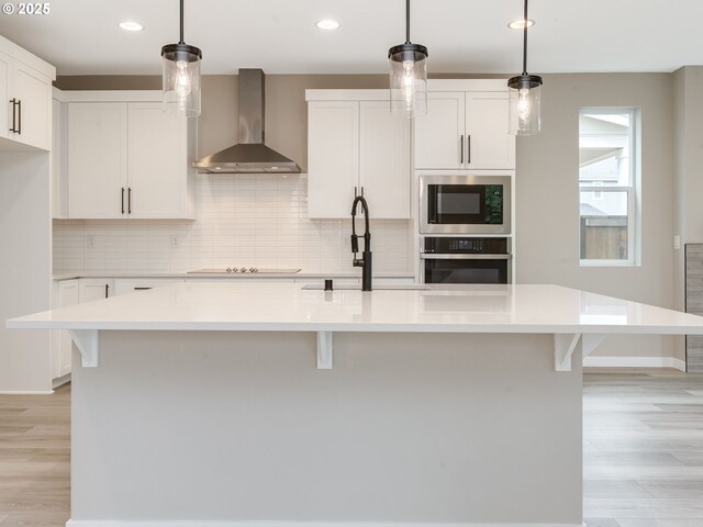 kitchen featuring tasteful backsplash, wall chimney range hood, appliances with stainless steel finishes, white cabinetry, and a center island with sink