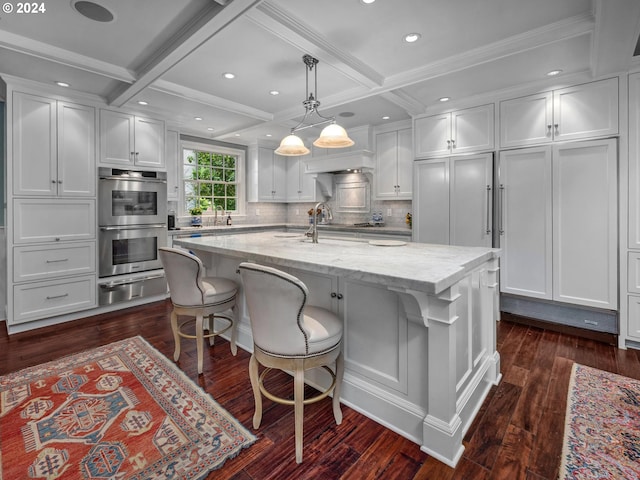 kitchen featuring white cabinets, coffered ceiling, a kitchen island with sink, and light stone countertops