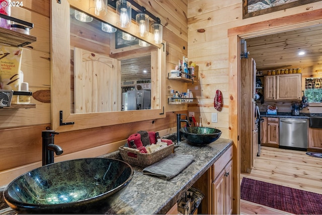 kitchen with sink, stainless steel dishwasher, dark stone countertops, wood walls, and light hardwood / wood-style floors