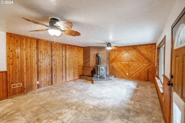 unfurnished living room featuring a wood stove, wood walls, ceiling fan, and a textured ceiling