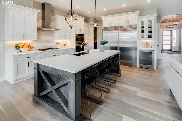 kitchen featuring light hardwood / wood-style flooring, tasteful backsplash, stainless steel appliances, a center island with sink, and wall chimney range hood