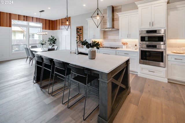 kitchen featuring a center island with sink, light wood-type flooring, backsplash, wall chimney exhaust hood, and appliances with stainless steel finishes