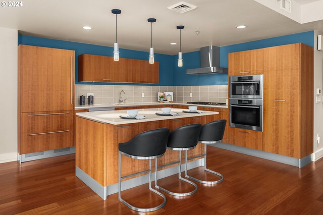 kitchen with tasteful backsplash, wall chimney range hood, hanging light fixtures, hardwood / wood-style floors, and a breakfast bar area