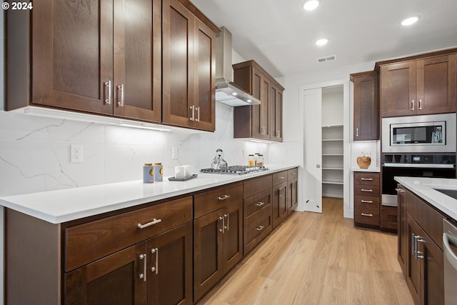 kitchen featuring appliances with stainless steel finishes, wall chimney exhaust hood, tasteful backsplash, and light hardwood / wood-style flooring