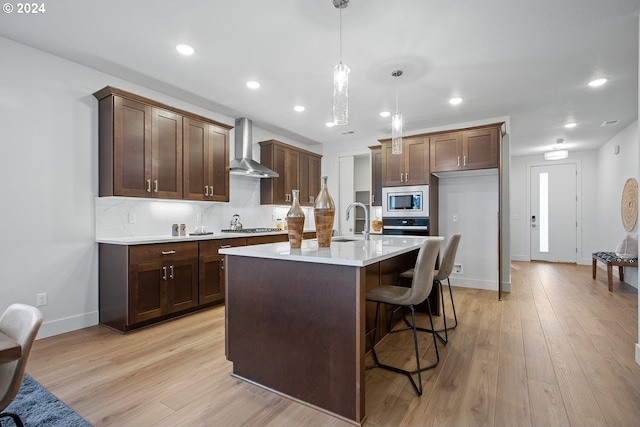 kitchen featuring pendant lighting, a center island with sink, light hardwood / wood-style flooring, wall chimney range hood, and stainless steel appliances