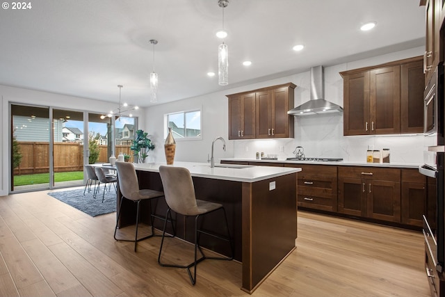 kitchen featuring pendant lighting, sink, an island with sink, wall chimney range hood, and light hardwood / wood-style flooring