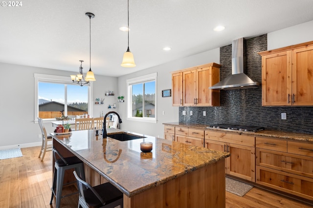 kitchen with wall chimney range hood, sink, light wood-type flooring, stainless steel gas cooktop, and a kitchen island with sink