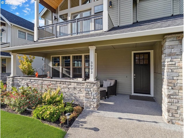 entrance to property with stone siding, covered porch, a shingled roof, and a balcony