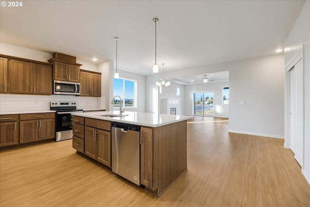 kitchen with sink, light wood-type flooring, an island with sink, hanging light fixtures, and stainless steel appliances