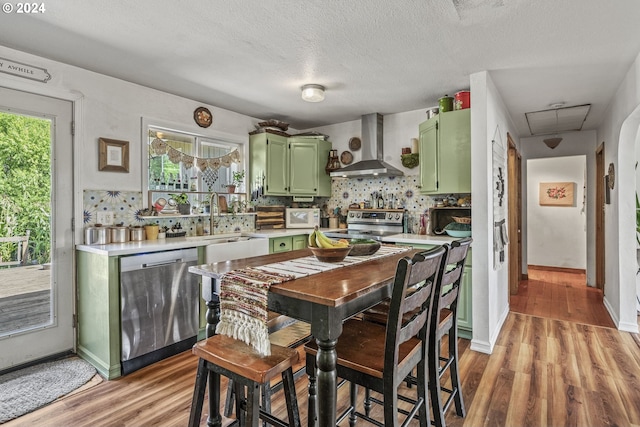 kitchen with backsplash, green cabinetry, appliances with stainless steel finishes, and wall chimney exhaust hood