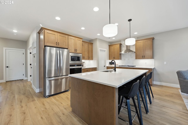 kitchen featuring hanging light fixtures, light wood-type flooring, an island with sink, stainless steel appliances, and wall chimney range hood