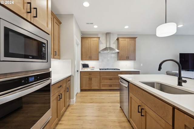 kitchen featuring sink, hanging light fixtures, stainless steel appliances, wall chimney range hood, and light wood-type flooring