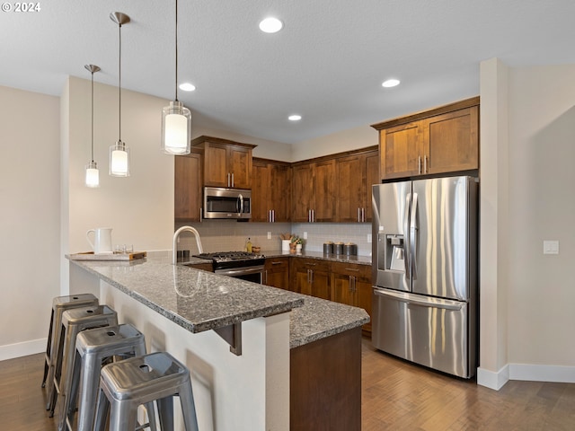 kitchen featuring dark hardwood / wood-style floors, kitchen peninsula, dark stone countertops, pendant lighting, and appliances with stainless steel finishes