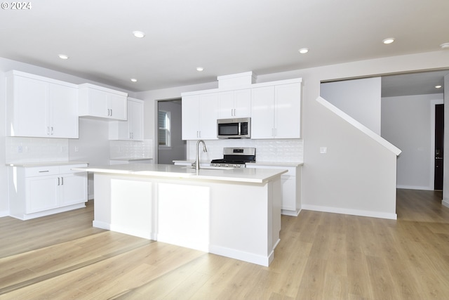 kitchen with an island with sink, stainless steel appliances, white cabinets, light wood finished floors, and light countertops