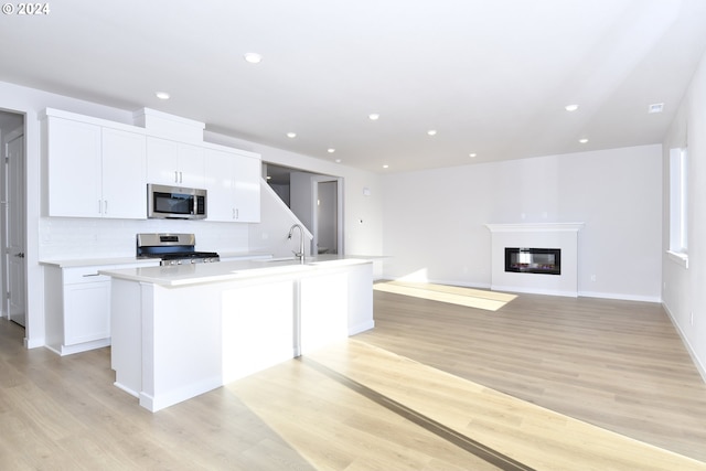 kitchen featuring a glass covered fireplace, a center island with sink, stainless steel appliances, and light wood-style flooring