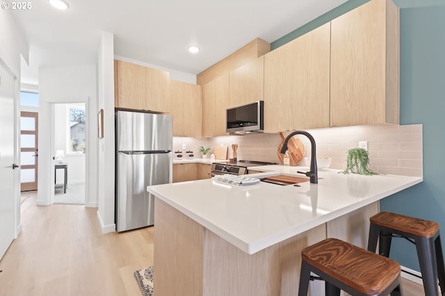 kitchen with a breakfast bar area, appliances with stainless steel finishes, backsplash, light brown cabinetry, and kitchen peninsula