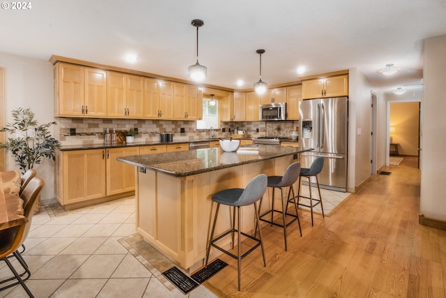 kitchen with appliances with stainless steel finishes, light brown cabinets, dark stone countertops, a center island, and hanging light fixtures