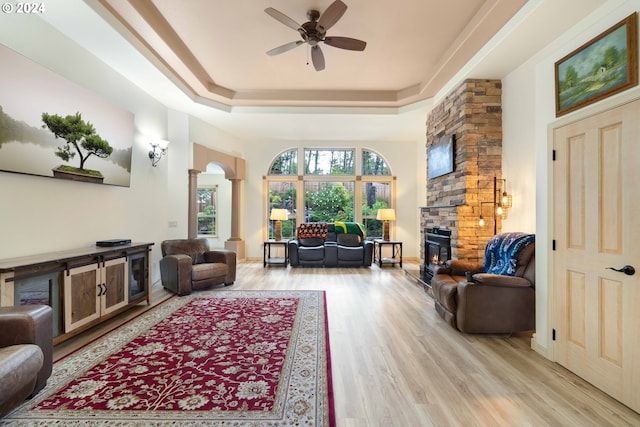 living room featuring ceiling fan, a raised ceiling, a stone fireplace, light hardwood / wood-style flooring, and decorative columns