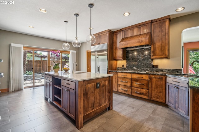 kitchen featuring stainless steel built in refrigerator, dark stone counters, a kitchen island with sink, and a healthy amount of sunlight