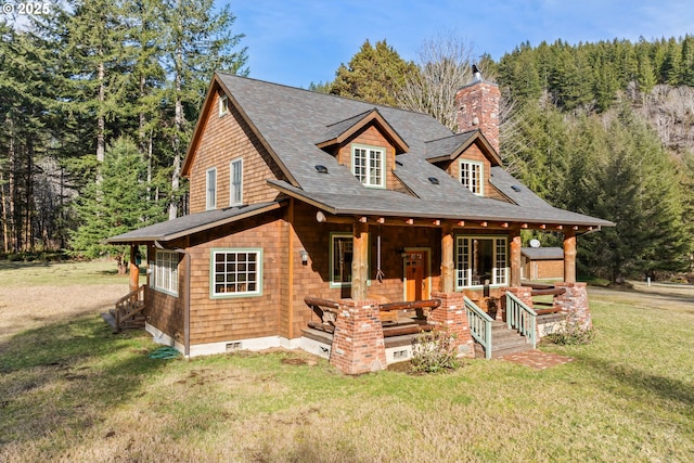 view of front of property with a forest view, roof with shingles, a porch, a chimney, and a front lawn