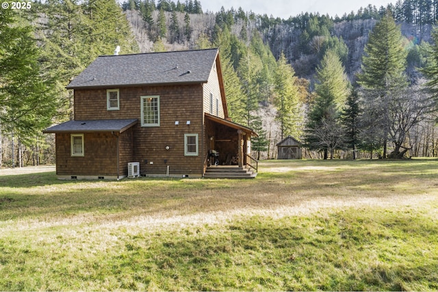 rear view of property with a yard, an outdoor structure, roof with shingles, and a wooded view